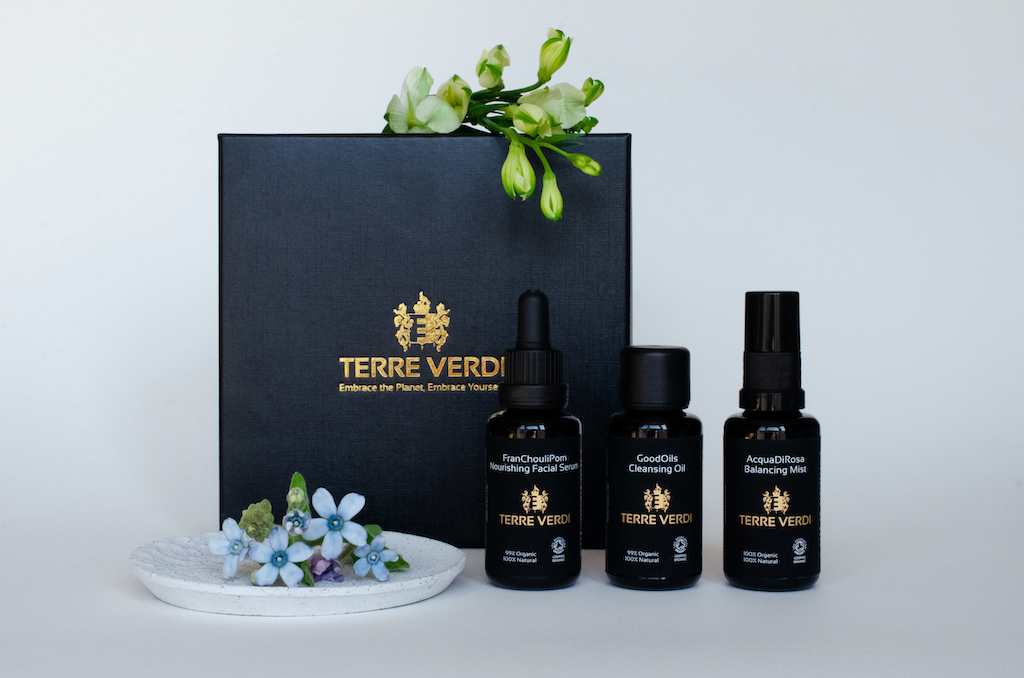 Terre Verdi Organic Gift Set for Face. Natural skincare gift set. All of the products are in black bottles and stand next to a luxury black box embossed in gold with the Terre Verdi logo. There are little blue flowers in front of the box and fresh green buds draped above.