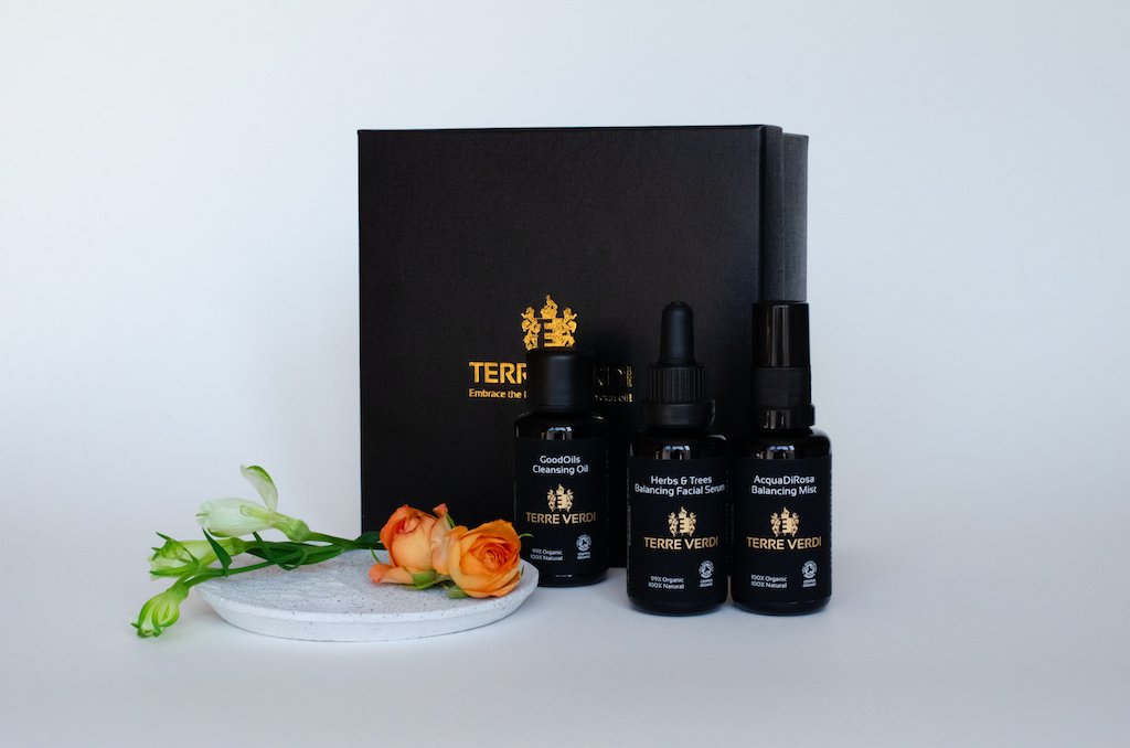 Terre Verdi Organic Gift Set for Face. Natural skincare gift set. All of the products are in black bottles and stand next to a luxury black box embossed in gold with the Terre Verdi logo. There are green flower buds and orange roses on a plate in front of the box.