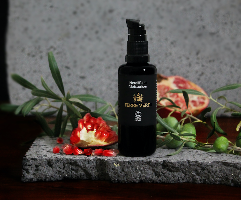 Terre Verdi Nerolipom Moisturiser. Certified organic face moisturiser. In a black glass bottle with a plastic pump. Bottle is sitting on a flat stone with a quarter of pomegranate next to it.