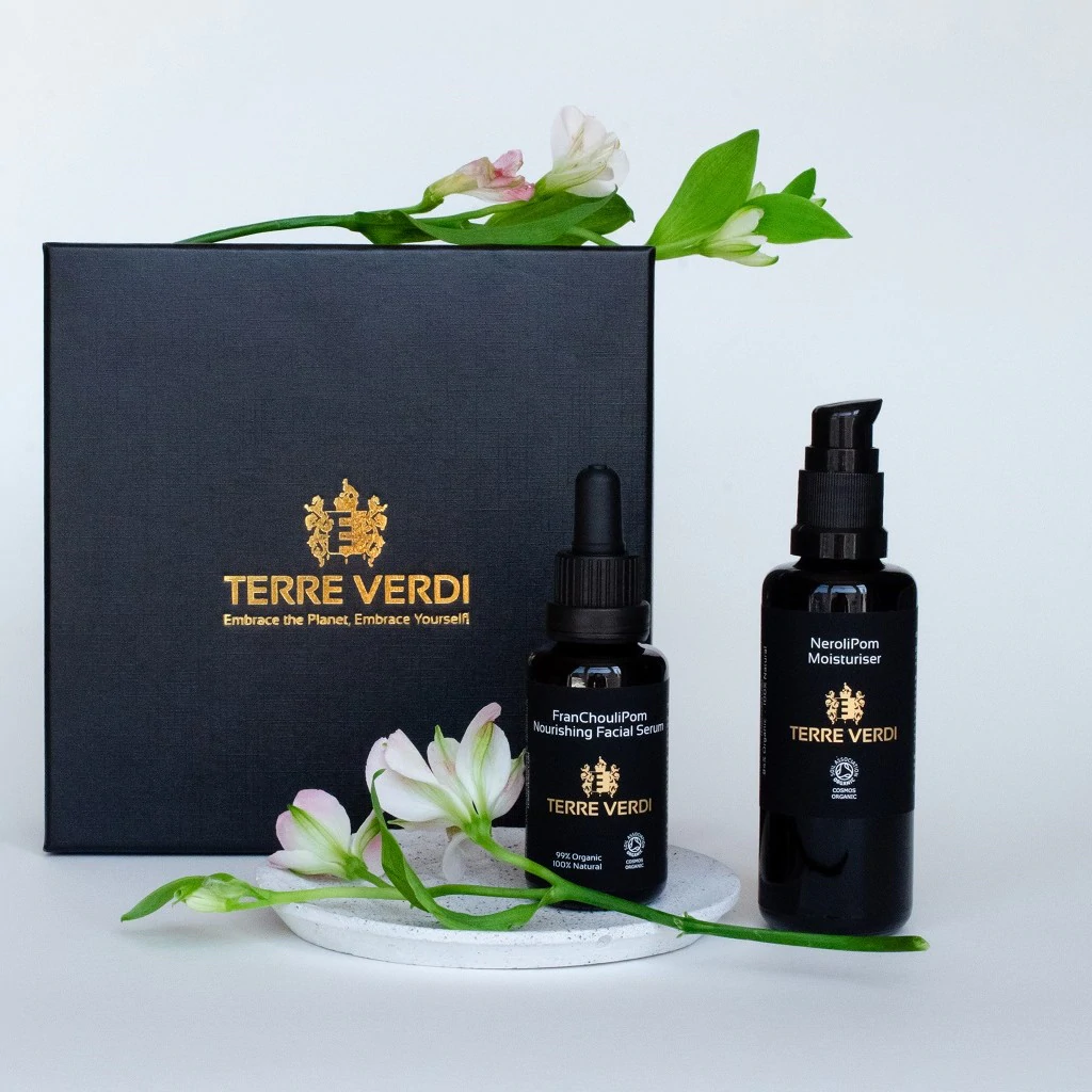 Terre Verdi Nourishing Gift Set. Certified organic skincare gift set. Both products are in black bottles and stand next to a luxury black box embossed in gold with the Terre Verdi logo. There are flowers draped in front of the products and on top of the box.