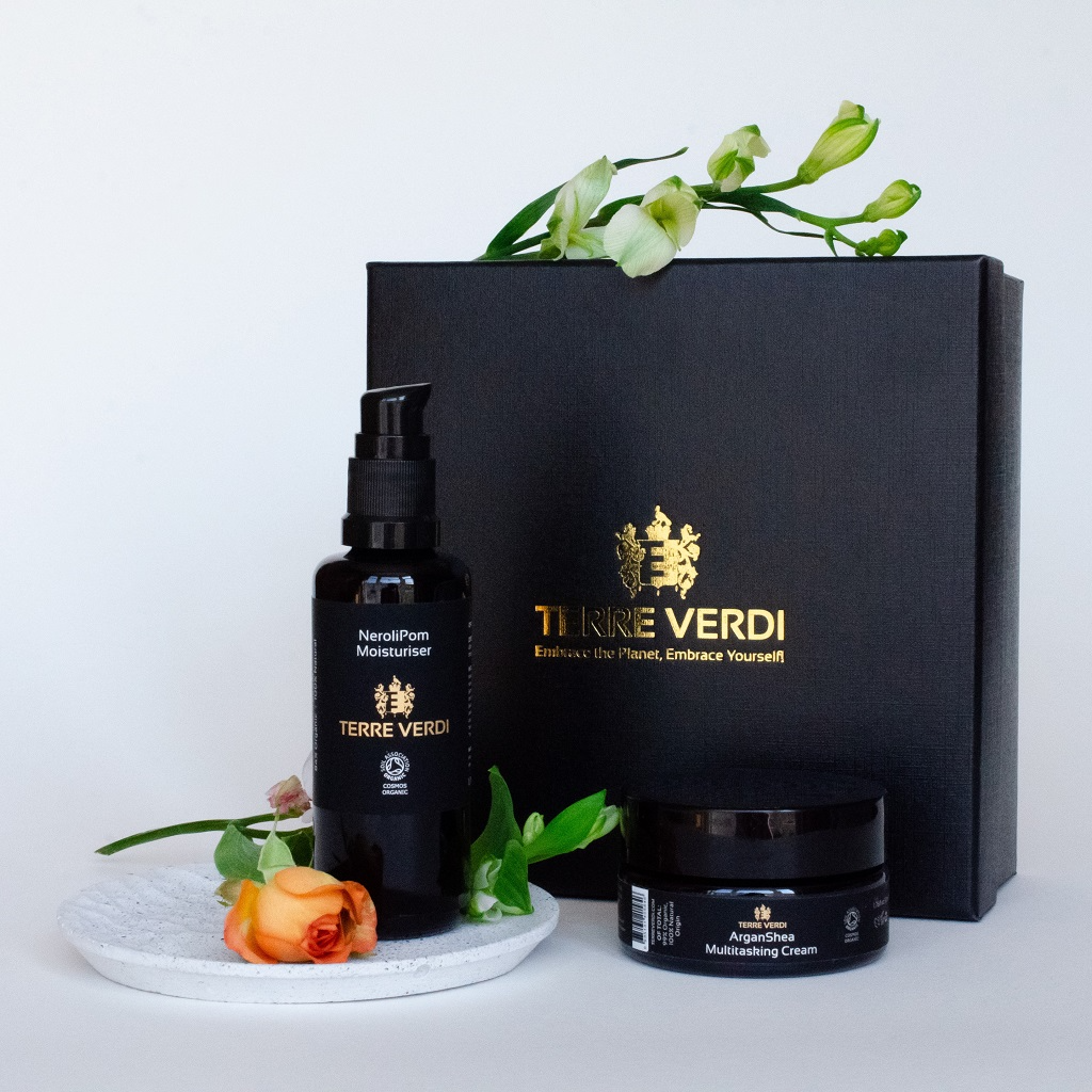 Terre Verdi Self Care Gift Set. Certified organic skincare gift set. Both products are in black bottles and stand next to a luxury black box embossed in gold with the Terre Verdi logo. There is a small orange rose in front of the moisturiser and green and white flower buds on top of the box.