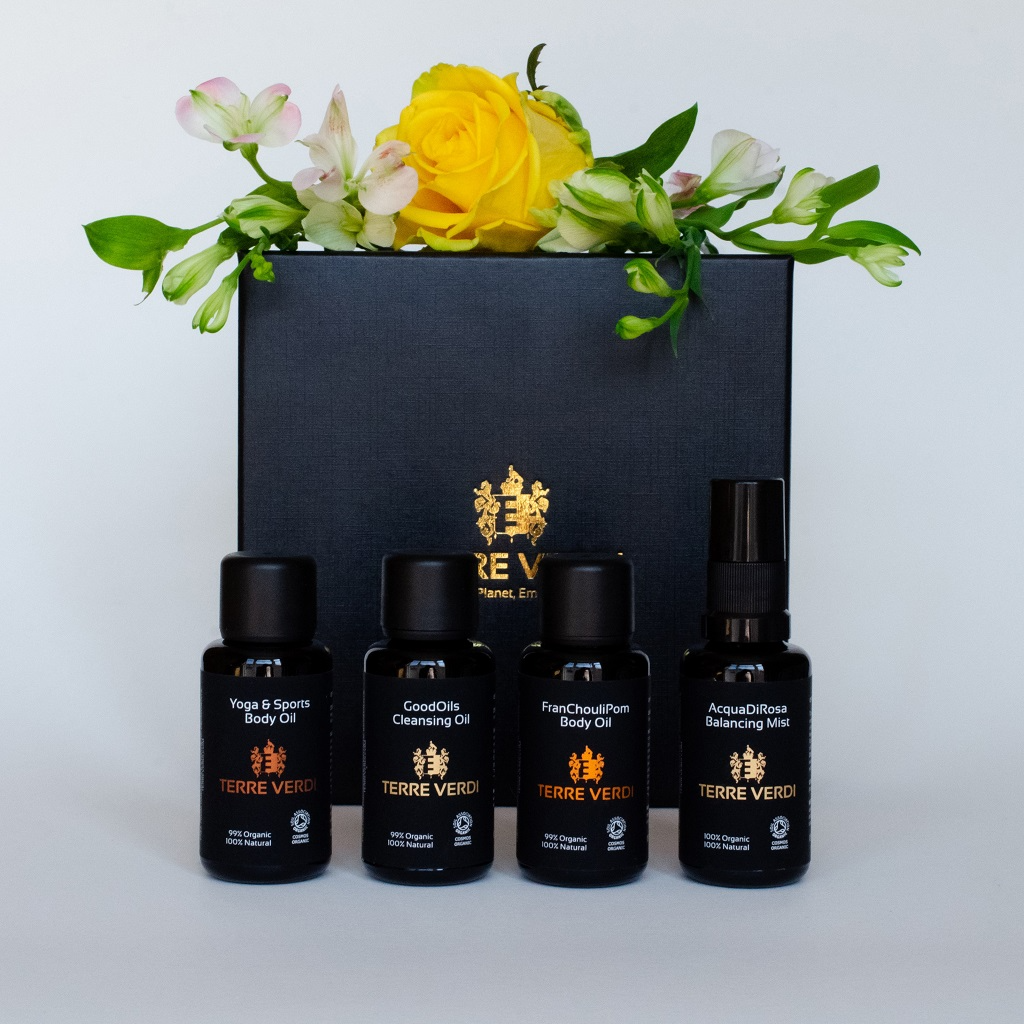 Terre Verdi Organic Travel Gift Set. Natural skincare gift set. All of the products are in black bottles and stand next to a luxury black box embossed in gold with the Terre Verdi logo. There is a small arrangement with a yellow rose and white flower buds on top of the box.