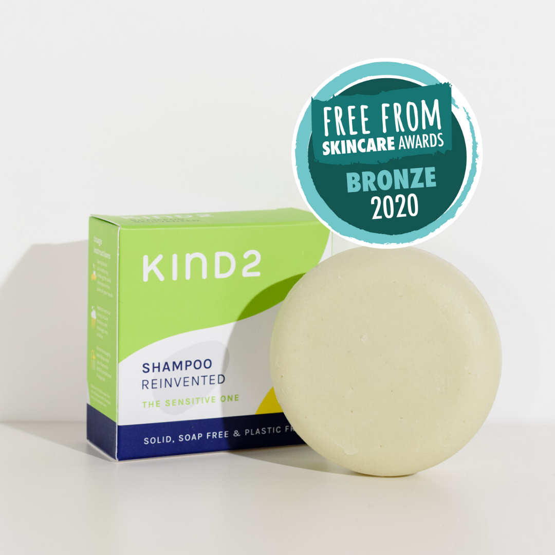 Round off-white plastic free KIND2 shampoo bar. Placed in front of its green, white and navy box. A Free From Skincare Shortlist Awards 2020 Winner Sticker on top of them.