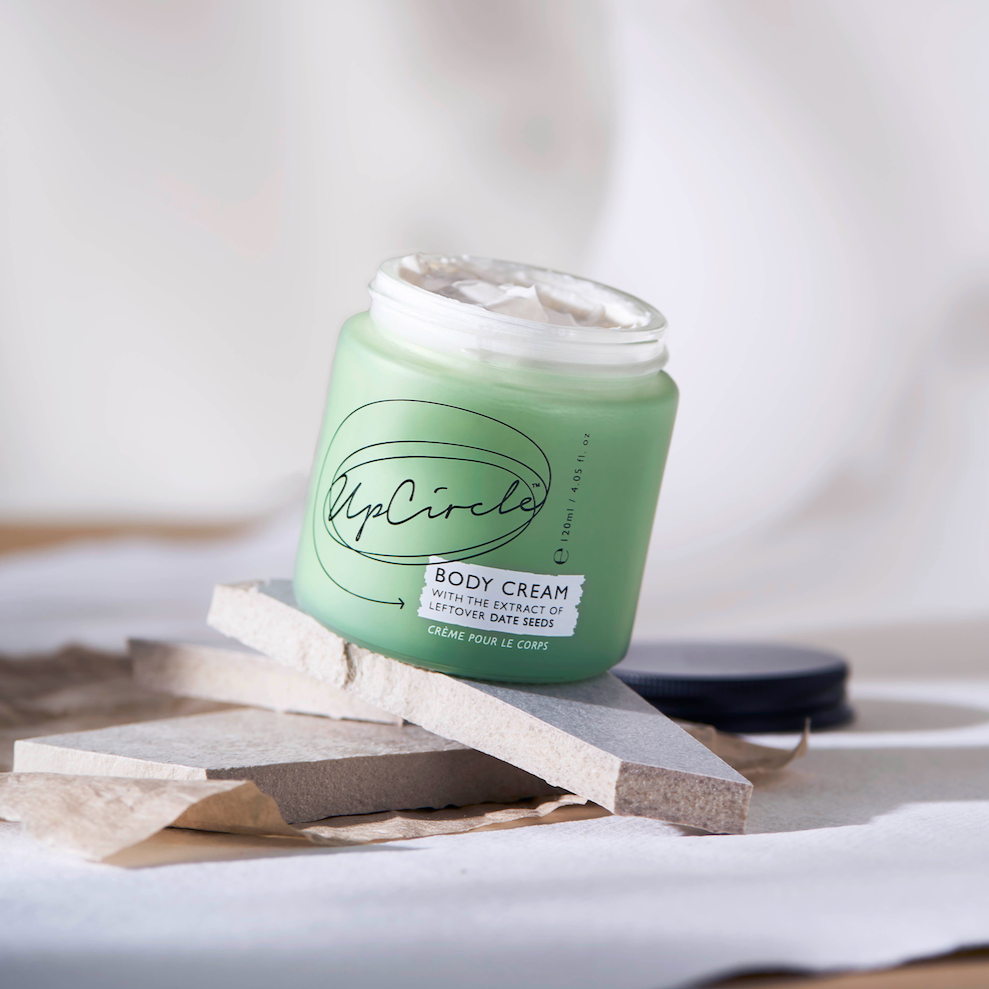 Upcircle Beauty Body Cream. Natural body moisturiser. The green glass jar is sitting open on some cuts of wood with a white sheet draped behind.