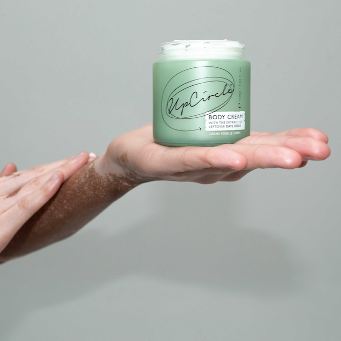 Upcircle Body Cream. Sustainable body lotion. The green glass jar is sitting open in a person's hand while they rub lotion into their arm.
