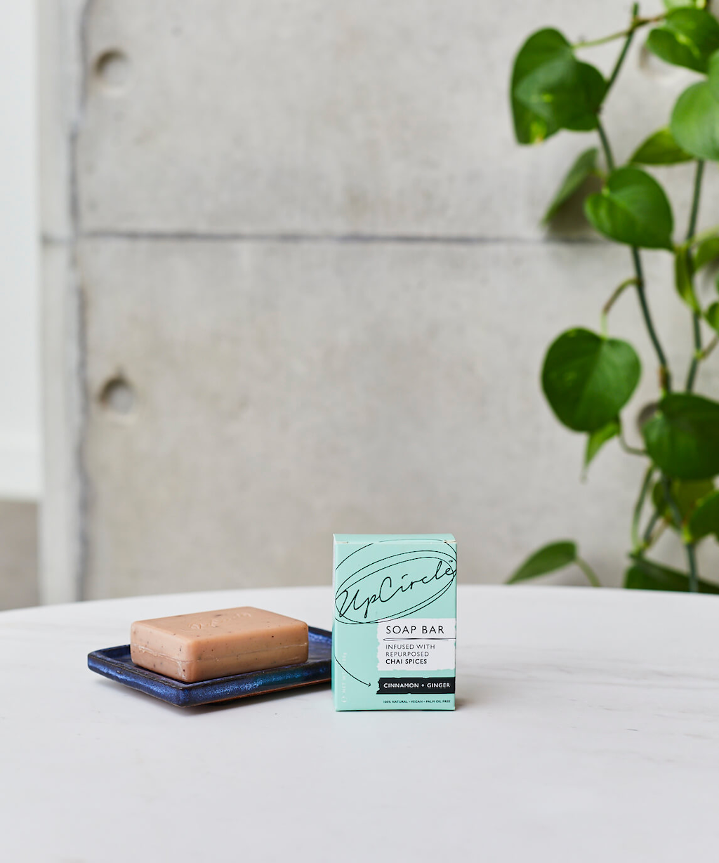 UpCircle Cleansing Soap Bar with Cinnamon and Ginger. Certified organic soap. The pink soap bar is sitting on a dark blue ceramic soap dish next to its green branded box on a white table with a concrete wall in the background.