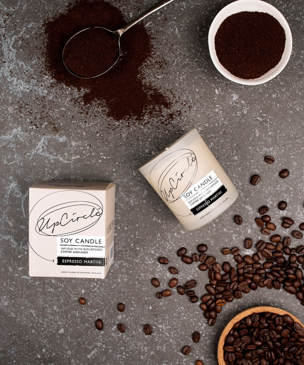 UpCircle Espresso Martini Soy Candle. Natural candle. The candle is shown laying on the ground next to it's box, with coffee beans and grounds dispersed around it.