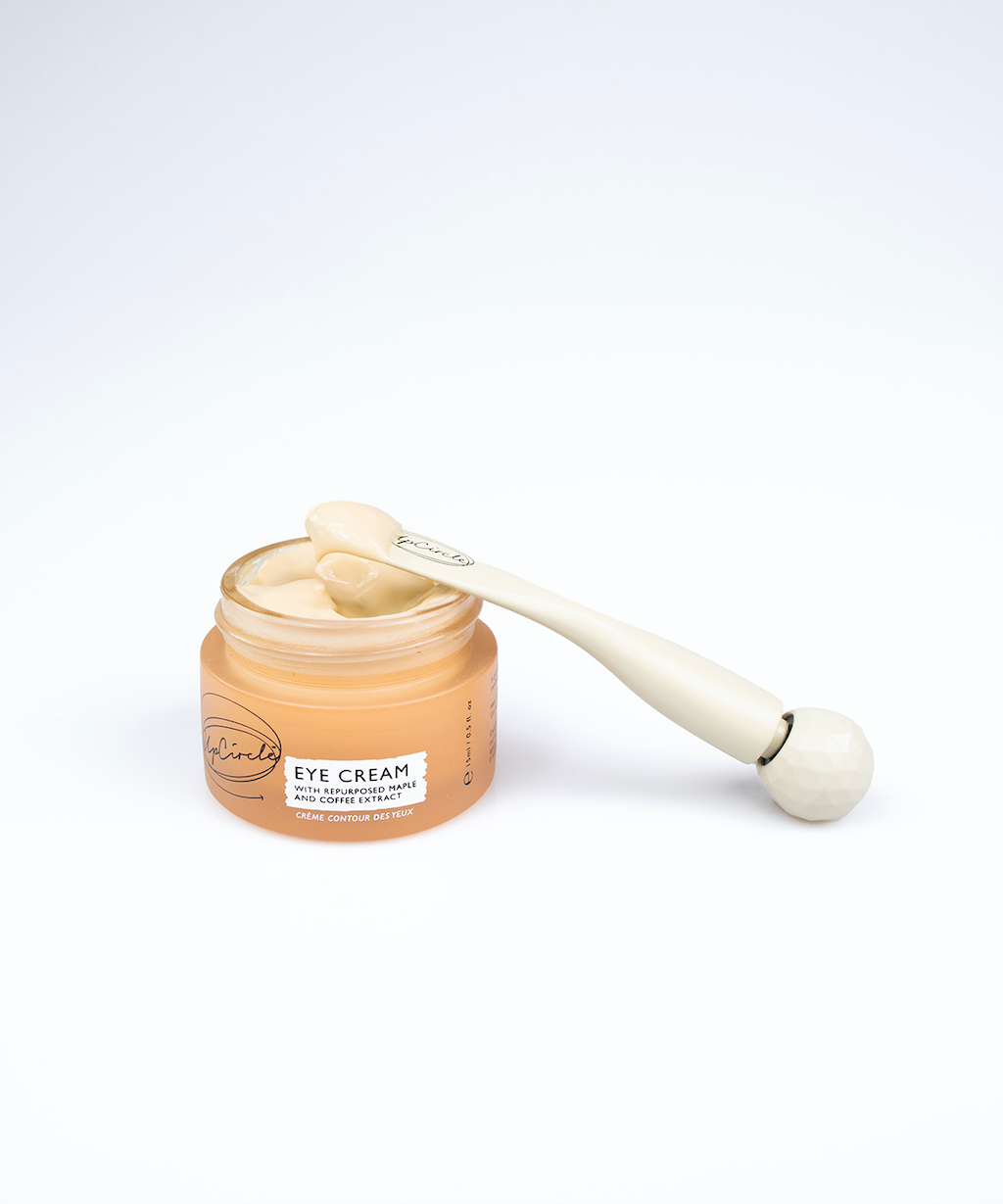 Eye roller tool for applying serums and eye creams can be seen next to a pot of upcircle eye cream which is open. the end of the eye roller is also a spatula so is shown with some of the mocha coloured cream on it. the cream is in an orange coloured glass pot