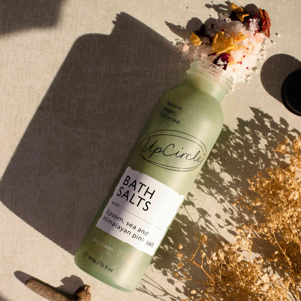 upcircle epsom, sea and himalayan pink bath salts can be seen spilling out of the translucent green glass bottle that's laid flat on a taupe fabric with dried flowers and wood surrounding it