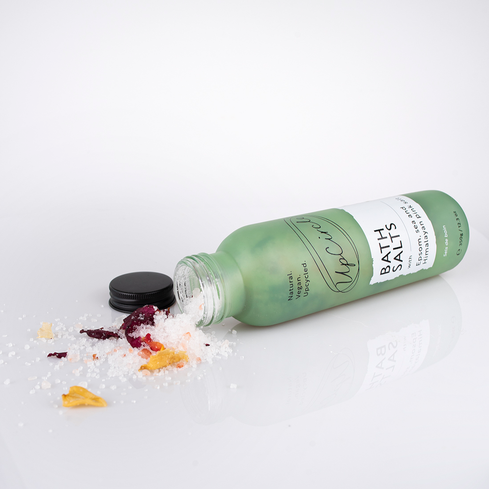white and pink bath salts with dried orange, yellow and dark pink dried flower petals can be seen spilling out of the light green glass bottle with black aluminium lid onto a reflective white surface