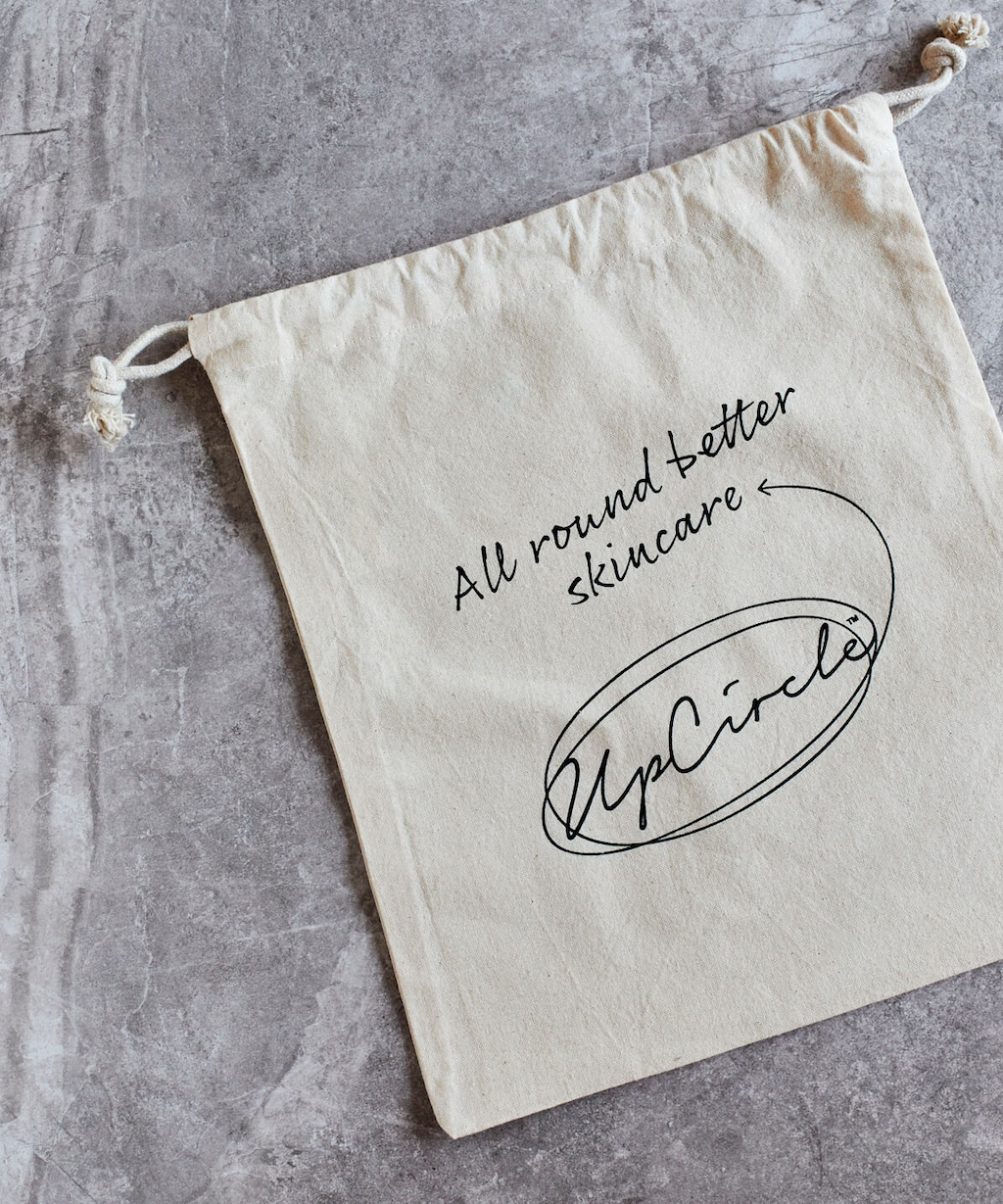 white linen bag with a drawstring which features the upcircle logo in a scrolling black font. Bag is on a grey textured surface and the text on the bag reads 'all round better skincare'