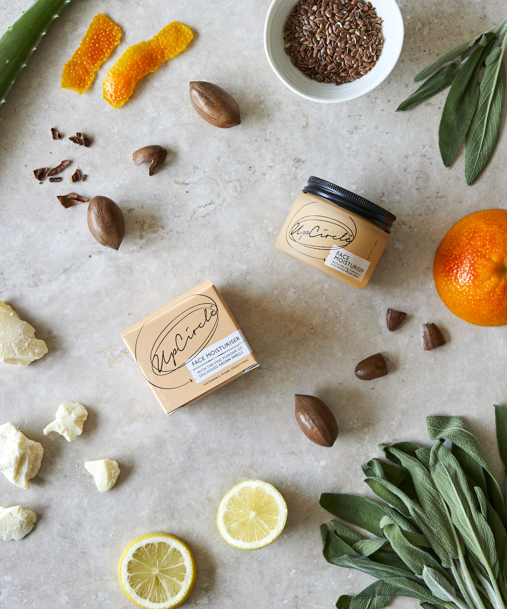 flat lay of upcircle's face moisturiser on a grey textured surface. featuring the orange glass and black aluminium lid, orange cardboard box and the raw ingredients within the moisturiser including sage, orange, shea butter, lemon and aloe vera