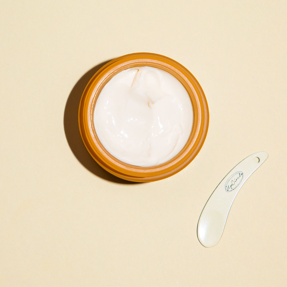 aerial shot of an open jar of upcircle moisturiser on a subtle orange background with a metal upcircle spatula next to it. you can see inside the orange glass jar with the texture of the moisturising cream inside