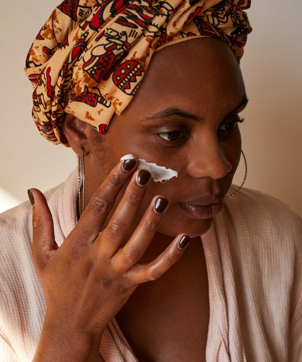 black woman in a blush pink robe and orange and red head scarf applying upcircle's face moisturiser