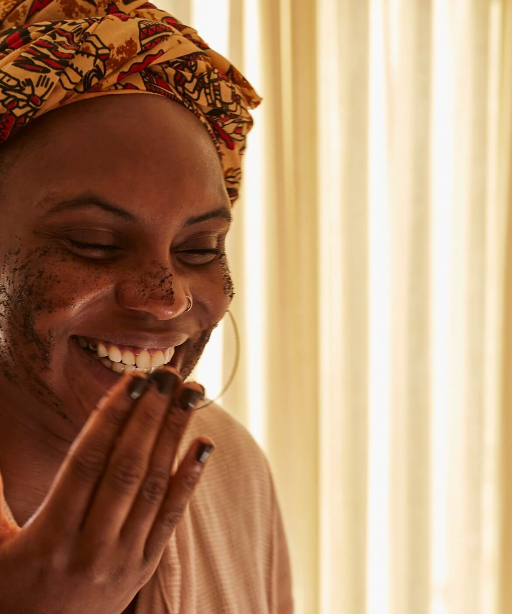 a black woman with an orange and red head scarf stands next to a window with light yellow curtains drawn. she is laughing and smiling as she applies the upcircle citrus and upcycled coffee grounds face scrub to her face
