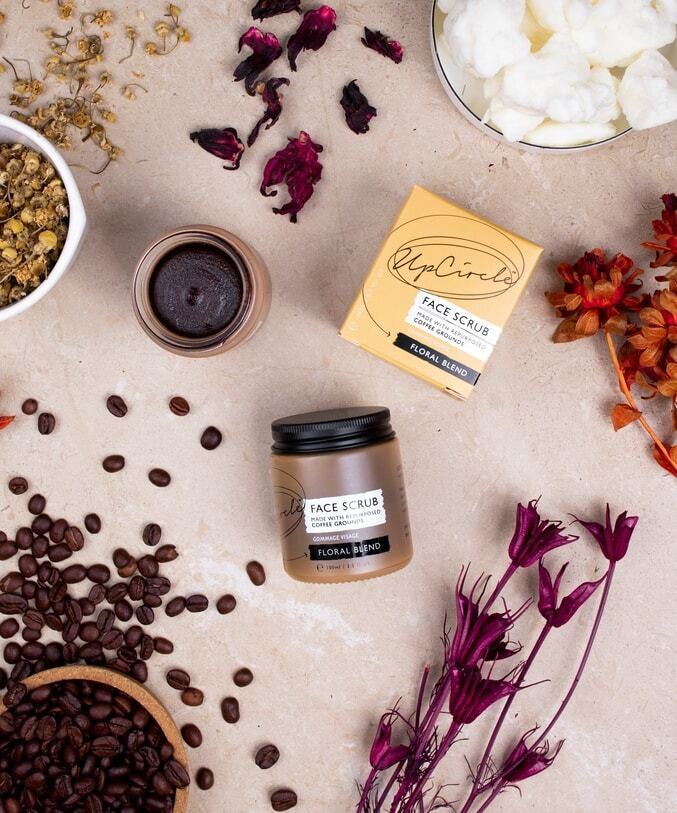 a jar of upcircle's floral and coffee face scrub can be seen surrounded by the natural ingredients it's made with like raw shea butter, coffee beans, chamomile flowers, hibiscus