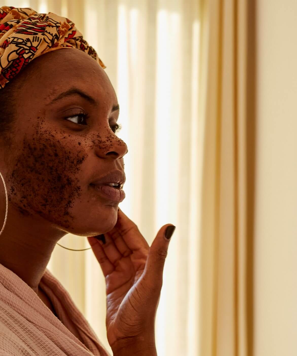 black woman wearing a red and yellow head scarf can be seen applying the coffee exfoliator to her face