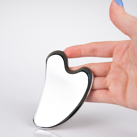 A hand with light skin and blue-painted nails holds a shiny, metallic Gua Sha tool with smooth, curved edges against a plain white background.