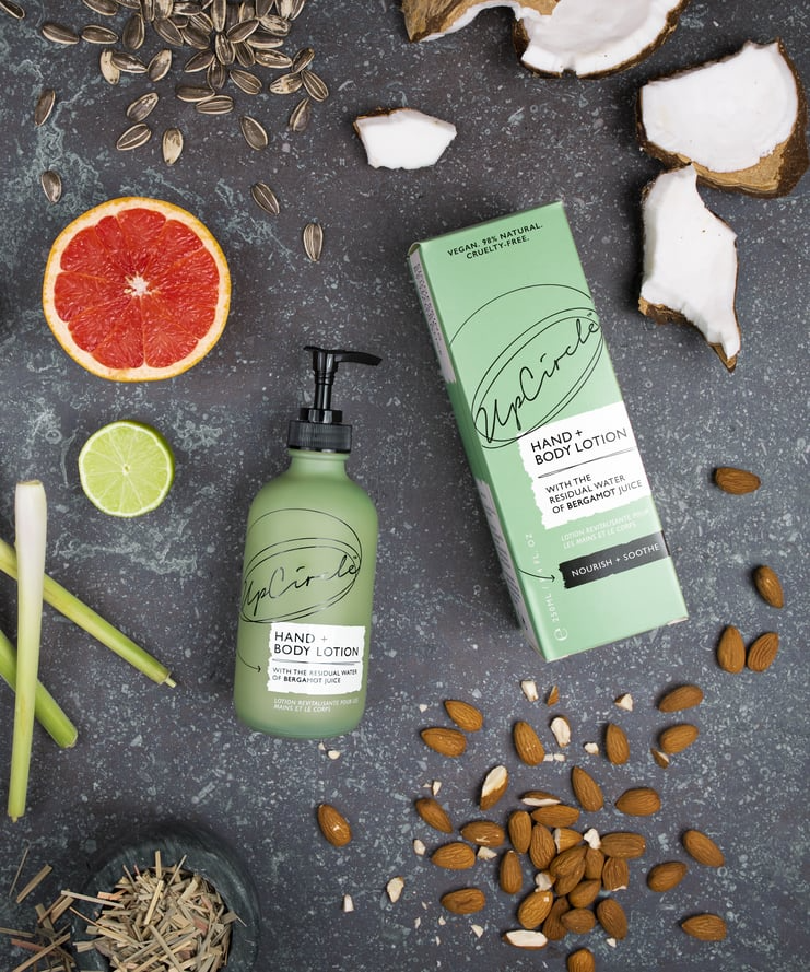 flat lay of the ingredients in upcircles bergamot hand lotion surrounded by the ingredients within the product. the background is a dark grey with lighter grey mottling and you can see the green glass bottle and cardboard box packaging of the hand cream surrounded by almonds, lemongrass, a lime and grapefruit sliced in half, sunflower seeds and a coconut that's been broken open