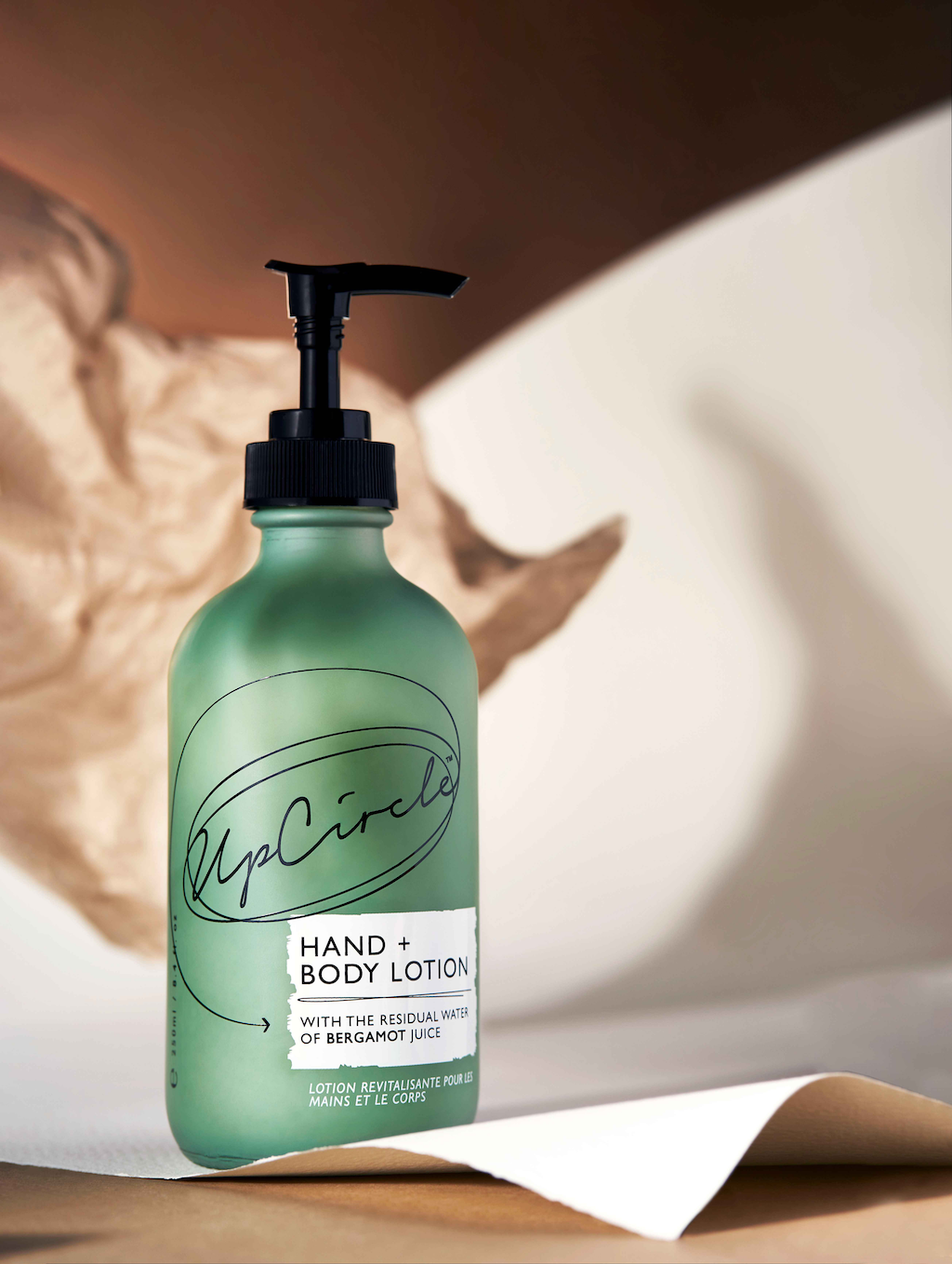 upcircle hand and body lotion lifestyle image where the frosted green glass pump bottle is bathed is gentle sunlight and sitting on top of a thick white card that's curling upwards