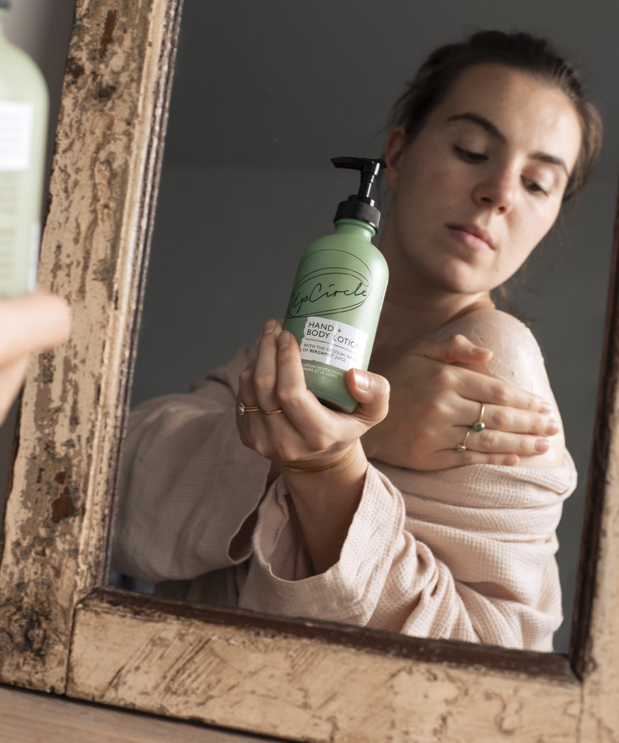 white woman with dark brown hair wearing a natural pink coloured top is looking into a mirror and applying upcircle's hand and body lotion to her exposed shoulder