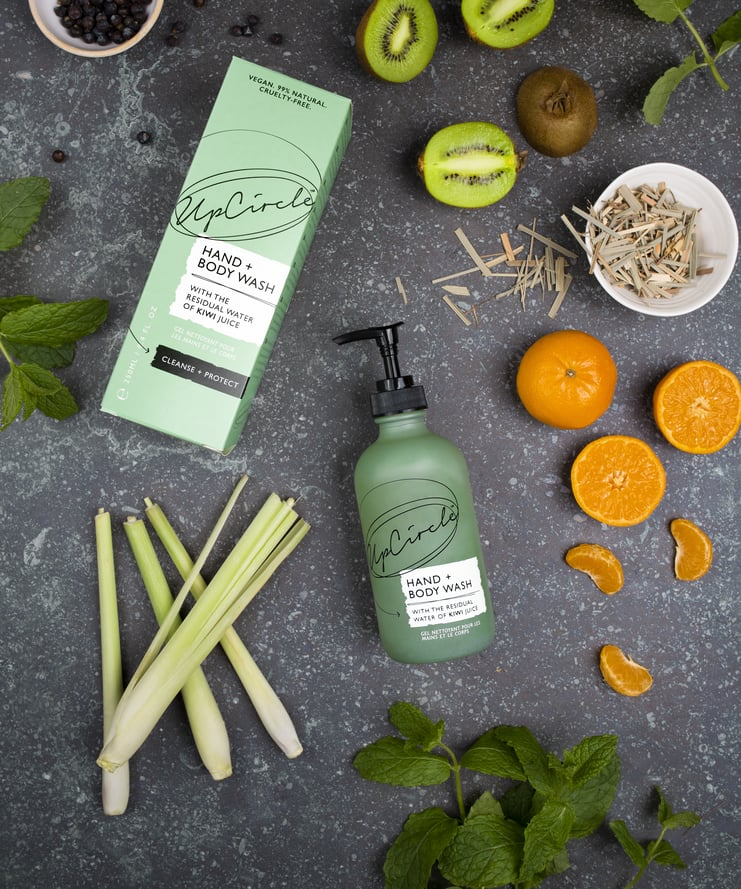 flat lay of the ingredients within the upcircle hand and body wash on a dark mottled grey background. the green glass bottle and cardboard box are surrounded by fresh mint, lemongrass, a whole, half and segmented orange, halved kiwi fruits 