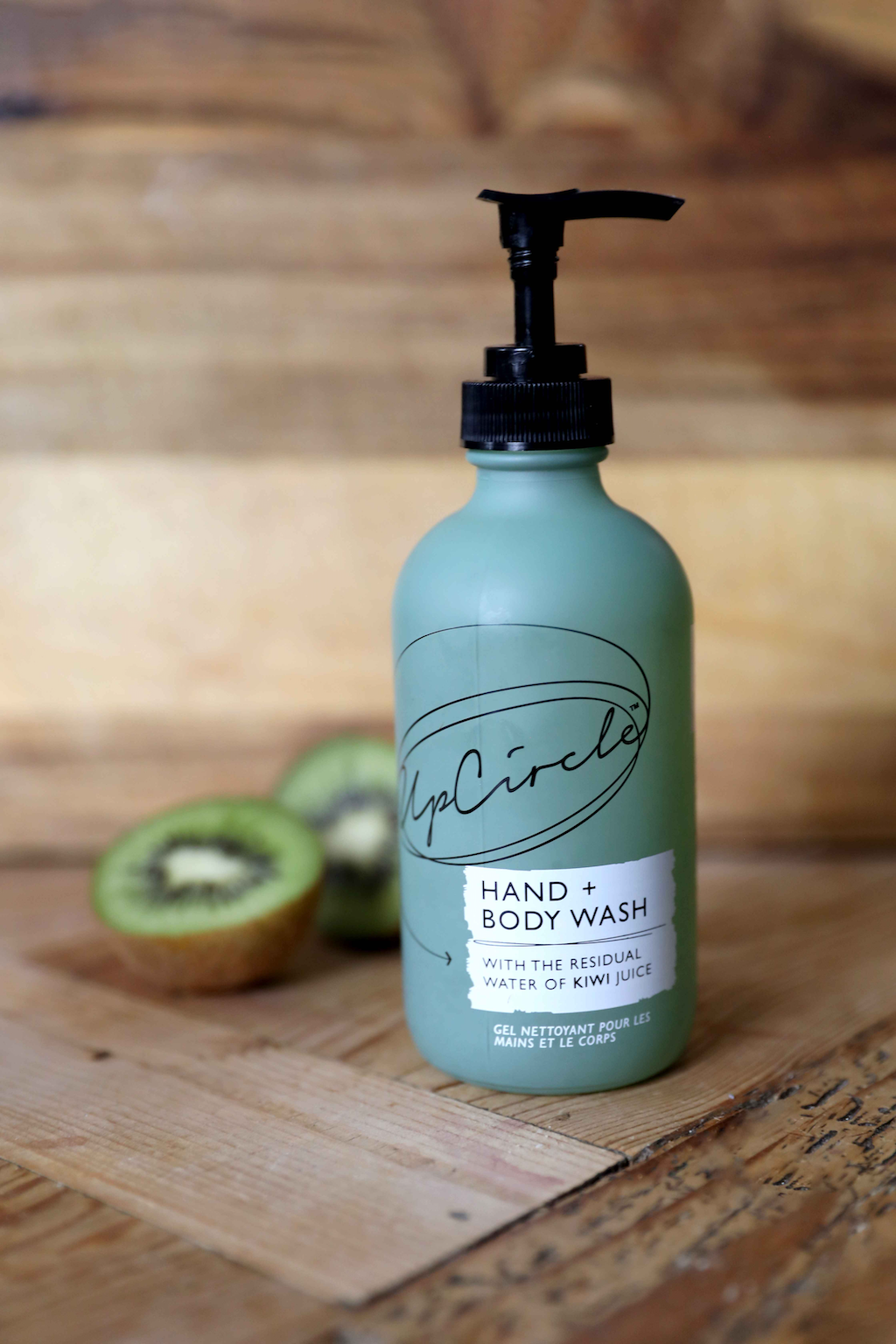 upcircle kiwi hand and body wash lifestyle image with the green glass bottle and black plastic pump sitting on a wooden board with a halved kiwi fruit in the background