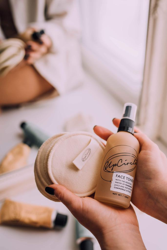 lifestyle image of the upcircle make up pads which can be seen being held in a white woman's hands alongside the orange glass bottle of the upcircle toner. in the background which is blurred you can make up some aluminium tubes of products and that the woman is sitting on a window ledge in the reflection in the mirror