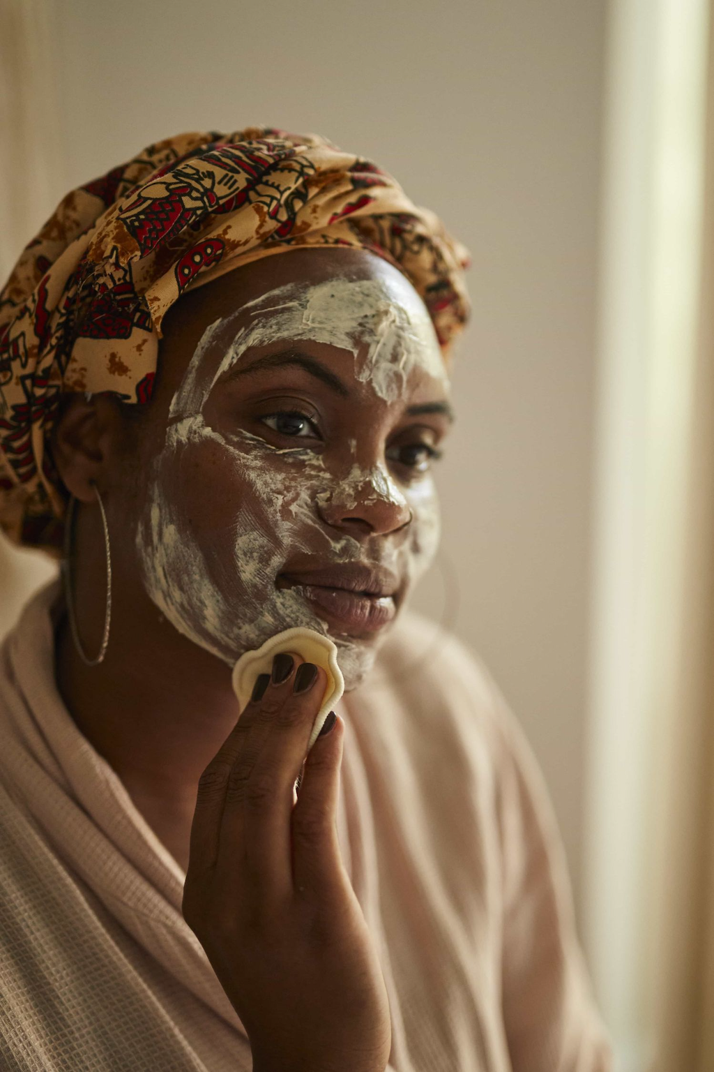 woman with brown skin and wearing a yellow, red and green head scarf with silver hoop earrings can be seen removing a creamy cleanser using the upcircle make up rounds